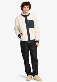 Fleecetrøje i creme med marineblå detaljer; har en høj krave, lynlås foran og sidelommer. Bæres med sorte jeans og beige sko.