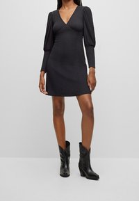 Robe noire avec un col en V et des manches longues bouffantes. Tissu texturé, silhouette ajustée, longueur au-dessus des genoux, associée à des bottines noires.