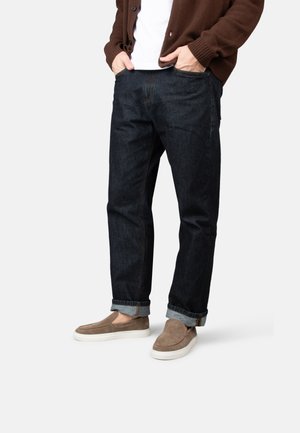 Mann trägt dunkle hochgekrempelte Jeans, braune Slip-On-Schuhe, weißes Hemd und braune Strickjacke, steht mit Händen in den Taschen vor weißem Hintergrund.