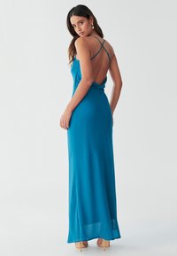 Maksi obleka iz zelenomodre tkanine z nabranim zadnjim delom. Ponaša se z tankimi čez hrbet prekrižanimi naramnicami in gladko, tekočo silhueto.