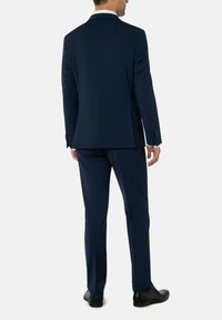 Man in een donkerblauw kostuum met zwarte schoenen, staand met de rug naar ons gekeerd tegen een lichte, effen achtergrond.