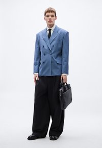 Sacou albastru cu dungi fine, cu două rânduri de nasturi, cămașă albă, cravată neagră, pantaloni negri largi și geantă neagră din piele, stând pe un fundal alb simplu.