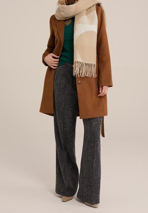 Femme portant un manteau marron, une écharpe beige et crème à motifs, un pull vert, un pantalon large gris et des chaussures beiges sur un fond uni.