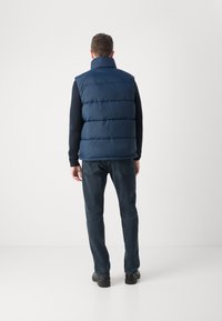 Marinblå pufferväst med horisontell vaddering, hög krage, bärs över en svart långärmad tröja, tillsammans med mörka jeans och svarta skor.