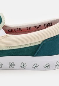 Zelené plátěné tenisky s bílou gumovou podrážkou a opakujícím se květinovým vzorem. Uvnitř růžové podšívky je text "NIKDY VE TMĚ."