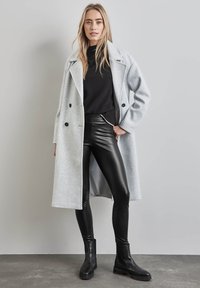 Grijze wollen combinatiejas met een dubbele rij knopen en zwarte knopen, gecombineerd met zwarte leerachtige leggings en zwarte enkellaarsjes.