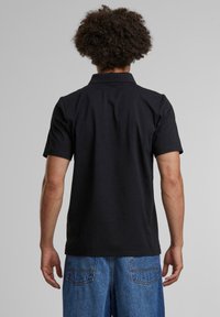 Svart kortermet polo-skjorte med krage, designet med en jevn tekstur. Båret sammen med blå denimjeans, som viser en enkel, ren silhuett.