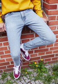 Jeans de mezclilla azul claro con un corte slim, con un sutil desvanecido y bordes desgastados. Combinados con zapatillas color burdeos y una sudadera amarilla.