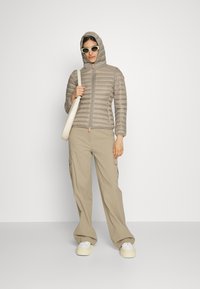 Lichtbeige pufferjack met capuchon, ritssluiting en een horizontaal gestikt patroon. Gecombineerd met beige cargobroek en witte sneakers.