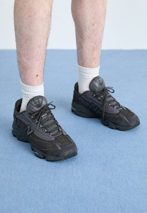 Beine mit weißen Socken und schwarzen New Balance 1000 Sneakers, die auf einem hellblauen Teppichboden stehen.