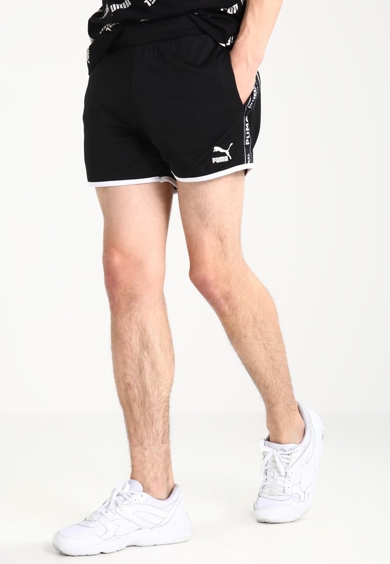 Shorts de sport noirs avec taille élastique, garnitures blanches et logo Puma sur le côté ; associés à des baskets blanches avec des semelles texturées.