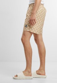 Beige fürdőnadrág fehér tengeri ló mintával, rugalmas derékkal és oldalsó logóval. Fehér, slip-on szandálokkal párosítva.
