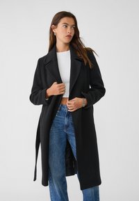 Svart ullblandad kappa med dubbelknäppning, slag och midjeband. Buren över en vit crop top och blå jeans.
