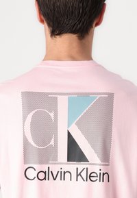 T-shirt en coton rose avec un design graphique au dos comportant "CK" en formes géométriques noires et bleues, ainsi que le texte "Calvin Klein".