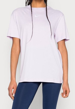 Kobieta nosząca luźną jasnofioletową koszulkę Nike i ciemnoniebieskie legginsy, stojąca na jednolitym tle.