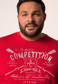T-shirt rossa in cotone con stampa grafica bianca di remi incrociati e la parola "COMPETITION." Il design include testo sul canottaggio e un aspetto vintage.