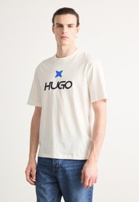 Cream cotton t-shirt with short sleeves, featuring a large black "HUGO" logo and a small blue star above it. Fitted design with straight hem.