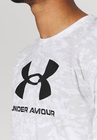 Weißes T-Shirt mit kurzen Ärmeln, das ein schwarzes Under Armour Logo zeigt, mit einem strukturierten, camouflagen Muster. Runder Ausschnitt.