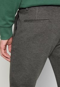 Pantalon de survêtement gris en tissu texturé, coupe slim, avec une poche arrière à rabat et une taille élastique, positionné sur un fond clair.