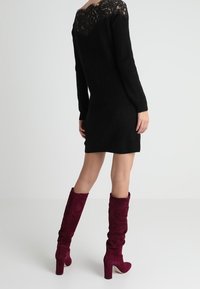 Robe noire en tricot avec des détails en dentelle au dos, associée à des bottes en daim bordeaux montantes avec un talon épais.