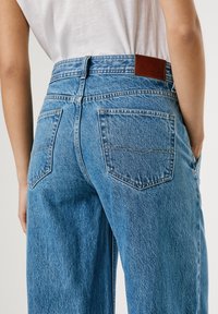 Jeans in denim blu a vita alta con una patch in pelle sul retro. Presentano due tasche posteriori con un delicato motivo di cucitura e una vestibilità comoda.