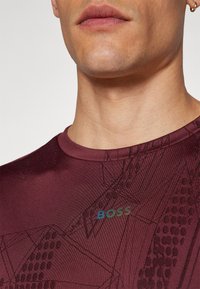Camiseta atlética burdeos con un patrón geométrico, cuello redondo y un sutil logo "BOSS" en colores degradados en el pecho.