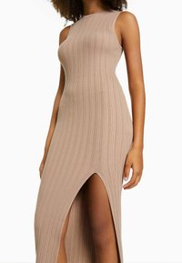 Robe côtelée longueur genoux en beige clair avec un col montant et des fentes latérales. Présente une silhouette ajustée et un design sans manches.