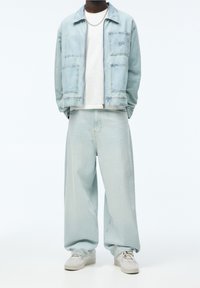 Světlá džínová bunda s náprsními kapsami a bílým tričkem, zkombinovaná s volnými světlými džínovými kalhotami a šedými teniskami. Součástí je i stříbrný řetízek jako doplněk.