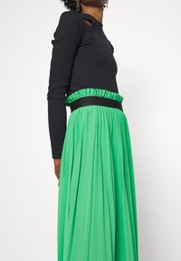 Haut noir à manches longues avec découpes, associé à une jupe maxi plissée verte comportant une taille froncée et une bande noire en accent.