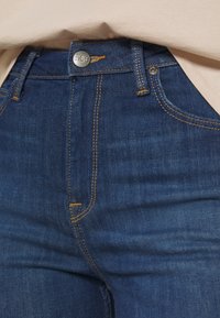 Gros plan sur un jean Lee bleu avec des surpiqûres jaunes, un bouton métallique, des passants de ceinture et une vue partielle d'une chemise beige rentrée au-dessus de la ceinture.