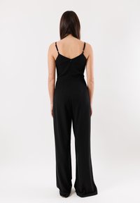 Zalio nero con un corpetto aderente e pantaloni a gamba larga. Presenta spalline regolabili e una zip sul retro con due tasche discrete. Tessuto liscio.