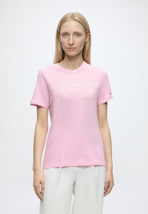 Jeune femme aux cheveux blonds raides porte un t-shirt rose Tommy Hilfiger et un pantalon blanc sur un fond clair uni.