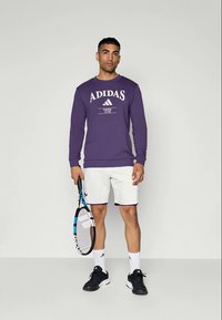 Lila pulóver fehér Adidas logóval, fehér rövidnadrággal és fekete sportcipővel párosítva. Teniszütőt tart a kezében.