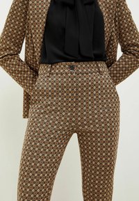 Kobieta w brązowej, wzorzystej marynarce oraz dopasowanych, wysokich spodniach, z czarną bluzką włożoną do środka, trzymająca ręce w kieszeniach marynarki.