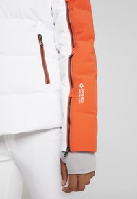 Gros plan sur la manche d'une veste blanche et orange avec plusieurs fermetures éclair noires et le logo "GORE-TEX INFINIUM" au-dessus d'un poignet gris.