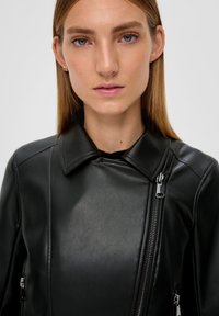 Veste en cuir noir avec un col, dotée de zips de chaque côté. Sa texture lisse et son design ajusté mettent en valeur son apparence élégante.