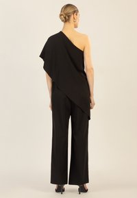 Asymmetrische zwarte jumpsuit met één schouder, wijduitlopende broekspijpen, zachte stof en een vloeiende stof die langs de rug valt.