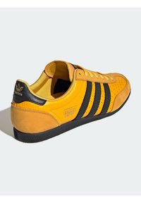 Žluté semišové tenisky s černými koženými pruhy, černou gumovou podrážkou a perforovanou špičkou. Na boku je logo "adidas Japan".