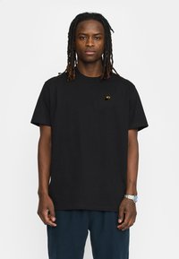 Zwarte katoenen t-shirt, korte mouwen, ronde hals, met een klein geel logo op de linkerkant van de borst. Draagt met donkerblauwe broek.