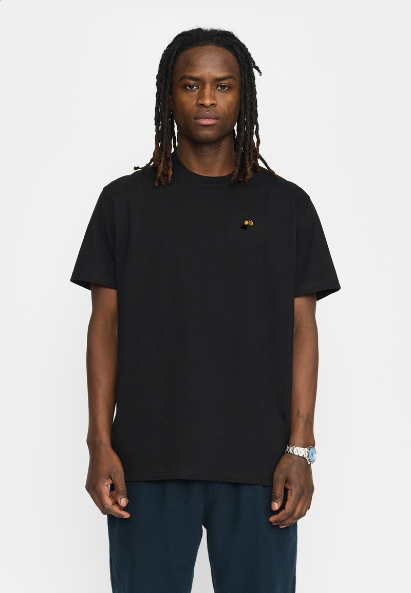 Zwarte katoenen t-shirt, korte mouwen, ronde hals, met een klein geel logo op de linkerkant van de borst. Draagt met donkerblauwe broek.