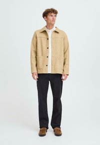 Beige wool blend jacket with a collar, front pockets, and black buttons, paired with a white shirt and dark pants. Brown suede shoes.