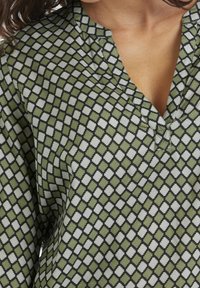 Grøn bluse med mønster i diamantmotiv i hvid og sort, med en let v-udskæring og diskret plissering ved halsen.