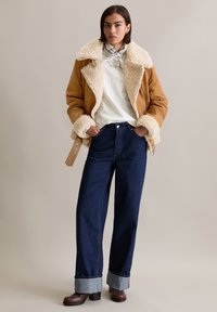 Bruine schapenvacht jas met een crème voering, gecombineerd met een witte shirt en donkerblauwe wijde jeans. Bruine laarzen met hak en een geprinte sjaal.