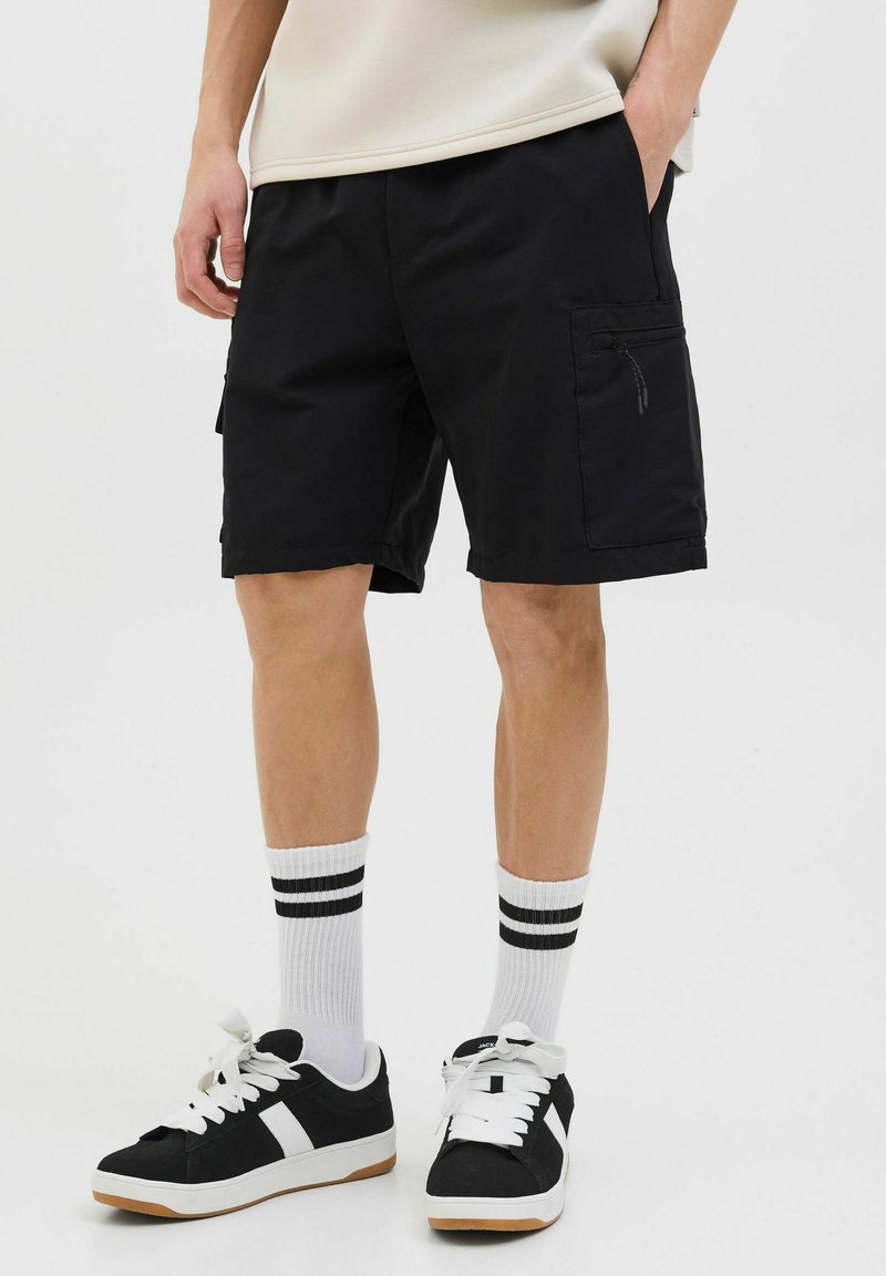 Schwarze Cargo-Shorts mit Seitentaschen, aus leichtem Stoff. Getragen mit schwarzen Sneakern und weißen Socken mit schwarzen Streifen.