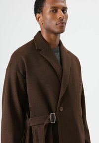 Bruine wollen jas met een reverskraag, knoopsluiting aan de voorkant en een riem met een zilveren gesp. Heeft een zachte textuur en een getailleerde pasvorm.