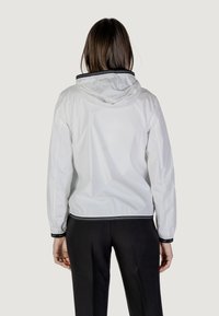 Giacca leggera bianca con cappuccio, con un bordo nero lungo il colletto e i polsini. Il tessuto ha una texture lucida e liscia.