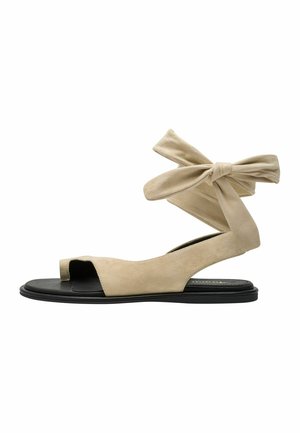 Beige flad sandal i ruskind med åben tå, sort sål og ankelrem bundet i en sløjfe.