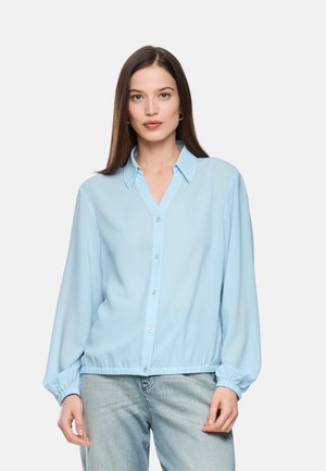 Frau trägt eine hellblaue, geknöpfte Bluse mit elastischen Bündchen und Bund, kombiniert mit hellblauen Jeans, steht vor einem einfarbigen Hintergrund.
