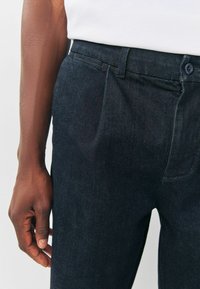 Dark blue denim pants feature a straight cut, subtle texture, a single button closure, and a side pocket; hand positioned on the thigh.