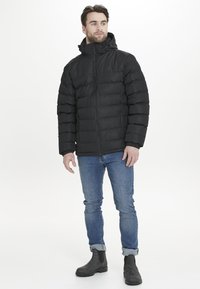 Chaqueta de plumas negra con cremallera frontal, capucha y textura a rayas. Lleva puesta sobre vaqueros azules y botas grises, de pie sobre un fondo blanco.